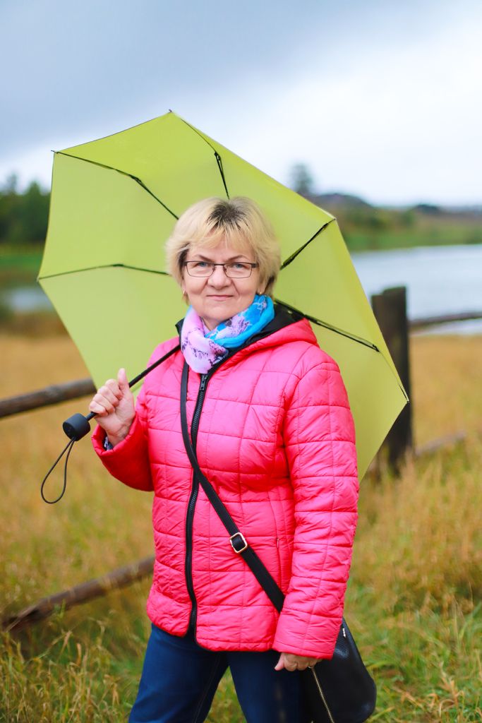 Natural Pharmaceuticals ImuBerin® Kobieta w różowej pikowanej kurtce trzyma jasnozielony parasol; spacer nad jeziorem w pochmurny dzień.