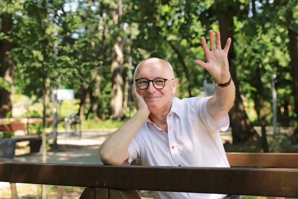 Natural Pharmaceuticals ImuBerin® Uśmiechnięty mężczyzna w okularach macha z ławki w parku, letni dzień wśród zieleni.