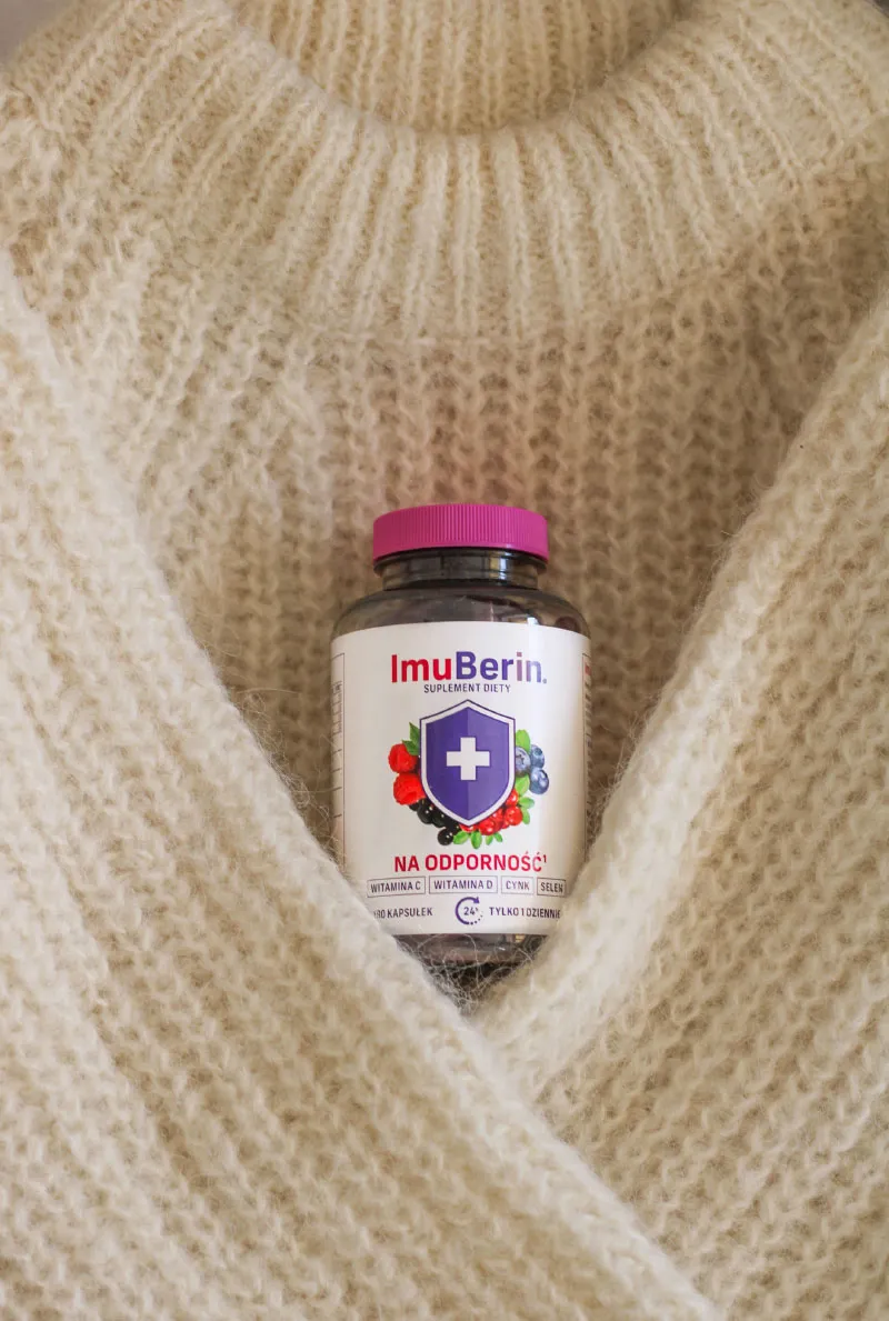 Natural Pharmaceuticals ImuBerin® Butelka suplementu ImuBerin „na odporność” z fioletową nakrętką wsunięta w miękki, kremowy sweter o grubym splocie.