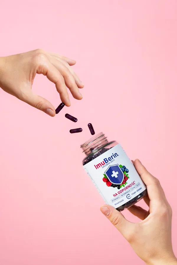 Natural Pharmaceuticals ImuBerin® Dwie dłonie i otwarty słoiczek ImuBerin®; fioletowe kapsułki spadają na różowym tle.