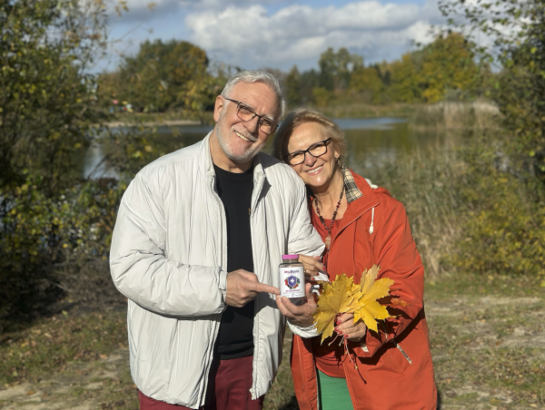 Natural Pharmaceuticals ImuBerin® Para seniorów nad jeziorem w jesiennej scenerii; uśmiechnięci, trzymają opakowanie ImuBerin i bukiet żółtych liści.